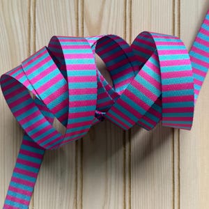 May include: A length of ribbon with alternating horizontal stripes of bright pink and turquoise blue. The ribbon is looped and curled, showcasing its pattern and texture. The background is a light-colored wooden surface.