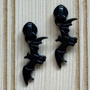 May include: Two black metal zipper pulls with a bat design. Each pull features a circular top with a small loop and a bat-shaped charm. The bats have outstretched wings and detailed features.