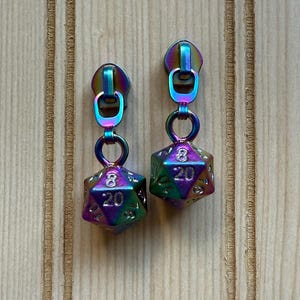 May include: Iridescent dice earrings. Each earring has a 20-sided die pendant, with the numbers 8 and 20 visible. The earrings are attached to iridescent zipper pull tops.