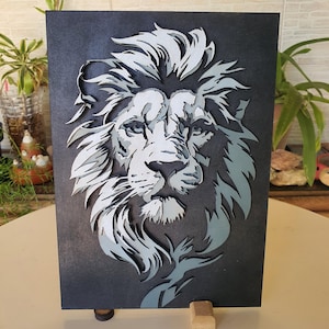 Puede incluir: Una obra de arte en papel en capas que representa la cabeza de un león. La obra utiliza tonos de gris y blanco sobre un fondo gris oscuro. La melena y la cara del león están intrincadamente detalladas, creando un efecto 3D. La pieza está montada sobre un soporte de madera.