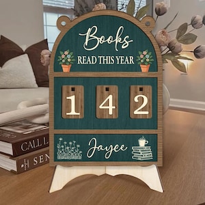 May include: Wooden book counter with the text "Books READ THIS YEAR" and the numbers "142". The counter has a teal background with a wooden frame and a stand. The name "Jaycee" is written on the bottom.