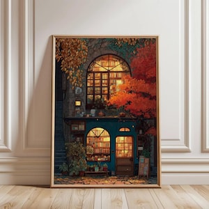 May include: A framed print of a cozy bookstore scene. The building has a teal facade, arched windows, and is surrounded by autumn foliage in shades of orange and gold. Bookshelves are visible through the windows, and the scene is warmly lit.