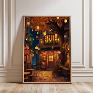 May include: Framed art print depicting a nighttime street scene with a restaurant. The artwork features warm lighting from lanterns and building windows, with a large tree and cobblestone street. The color palette includes warm oranges, browns, and blues.