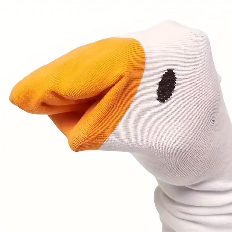May include: A white sock with a cartoon goose design. The sock features a bright orange beak and a black eye. The beak is textured, and the sock is made of a soft material.