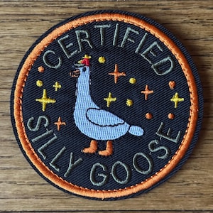 May include: Embroidered patch with a black background and an orange border. The patch features a cartoon goose with the words "CERTIFIED SILLY GOOSE" in white and orange. Yellow cross and dot accents are also included.
