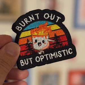 May include: Black patch with the text "BURNT OUT BUT OPTIMISTIC". The patch features a cartoon marshmallow on fire, set against a sunset-coloured background with trees. The patch is held by a hand.