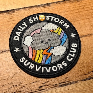May include: Circular embroidered patch with the text "DAILY SH*STORM SURVIVORS CLUB" in white on a black background. The design includes a grey cloud, rainbow, and lightning bolt. The patch is on a wooden surface.