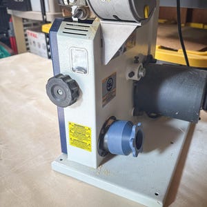 May include: A stationary power tool with a light gray base and a white and blue body. The tool has a black knob, a cylindrical dust collection port, and a blue attachment. Warning labels are visible.