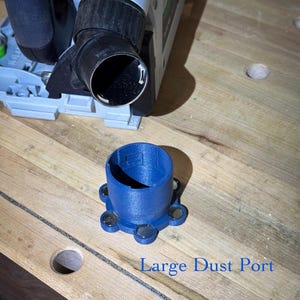 May include: A blue plastic dust port with a circular opening and a series of magnets on the base. The text "Large Dust Port" is printed below the port. A power tool is visible in the background, resting on a wooden surface.