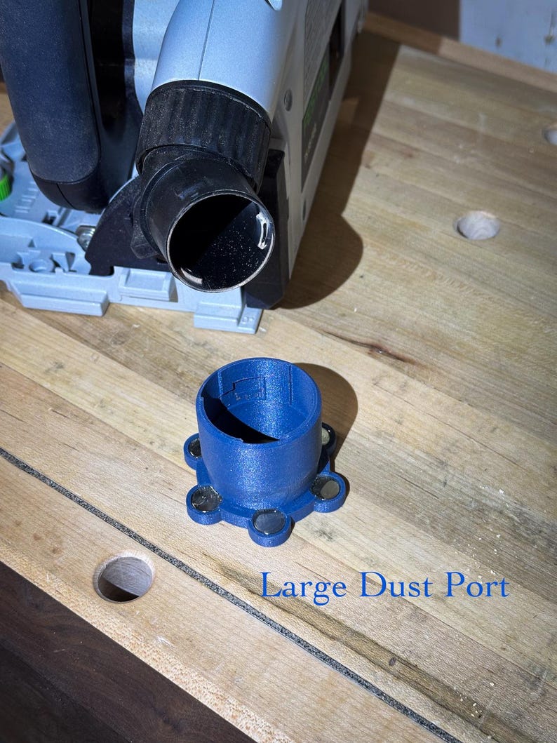 May include: A blue plastic dust port with a circular opening and a series of magnets on the base. The text "Large Dust Port" is printed below the port. A power tool is visible in the background, resting on a wooden surface.