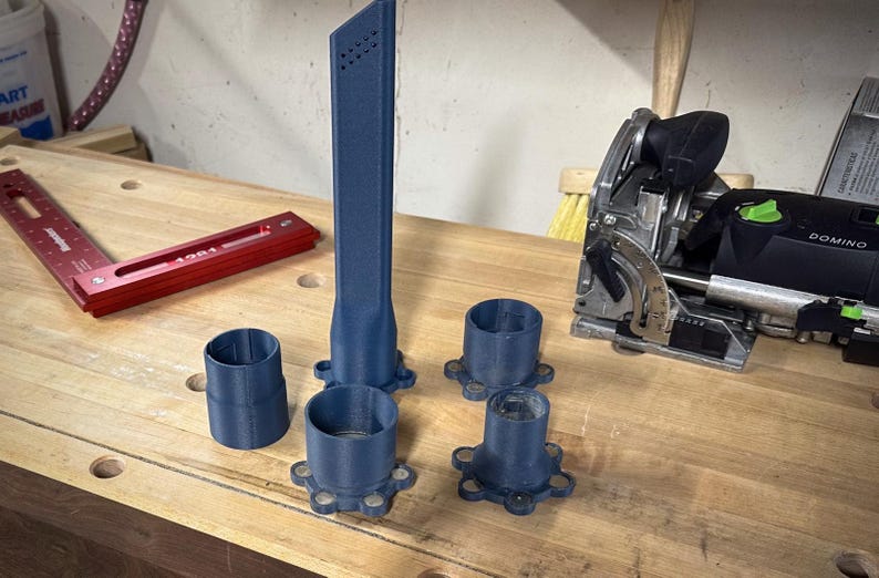 May include: A collection of blue 3D-printed woodworking tools and accessories. The set includes a tall, slender nozzle with air vents, several cylindrical attachments, and a red metal angle measuring tool. A black and green power tool is also visible.