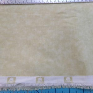 May include: Pale yellow fabric with a mottled pattern. A white border with a fringed edge and a repeating logo is at the bottom. A ruler is at the top, and a blue cutting mat is at the bottom.