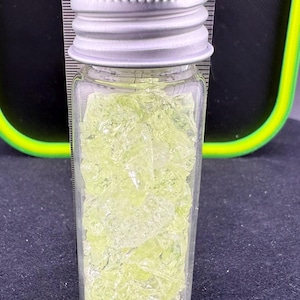 May include: A small, clear glass vial with a silver metal lid, filled with pale green, crystalline fragments. The vial is positioned vertically, with a ruler in the background showing measurements in inches.