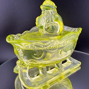 Puede incluir: Figura de Papá Noel de cristal amarillo verdoso en un trineo. La figura de Papá Noel está sentada, con barba larga y gorro. El trineo tiene adornos decorativos y patines. El cristal tiene calidad reflectante y el fondo es oscuro.