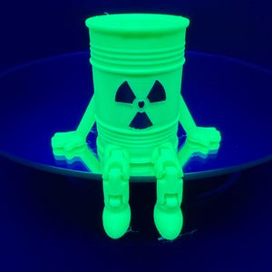 Può includere: Un barile verde neon in stile cartone animato con un simbolo di radiazione sul davanti. Il barile ha braccia e gambe, conferendogli un aspetto giocoso. Lo sfondo è blu scuro, creando un contrasto con la figura verde brillante.
