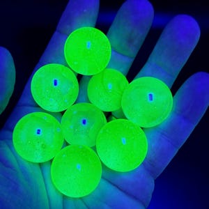 Puede incluir: Un conjunto de ocho canicas de vidrio esféricas y pequeñas que brillan con un color verde neón intenso bajo luz ultravioleta. Cada canica tiene una superficie lisa y translúcida, con una forma redonda uniforme y un efecto fluorescente.