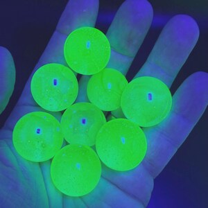 Puede incluir: Una colección de nueve esferas verdes brillantes con un centro azul, sostenidas en una mano. Las esferas son redondas y parecen estar hechas de un material translúcido. El fondo es azul oscuro.