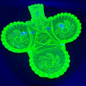 May include: A glowing green, three-part glass serving dish with a central handle. The dish has a scalloped edge and is decorated with etched floral and geometric patterns. The dish is illuminated with a black light, causing it to glow.