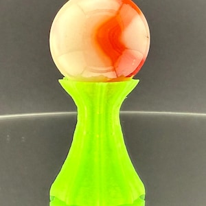 Könnte beinhalten: Eine dekorative Glaskugel mit roten und weißen Wirbeln, die auf einem hellgrünen Sockel ruht. Die Kugel ist kugelförmig und der Sockel hat ein geriffeltes Design.
