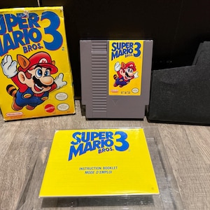 May include: A Super Mario Bros. 3 video game set. The yellow box features Mario in a tanooki suit. The set includes the game cartridge, instruction booklet, and a black storage case.