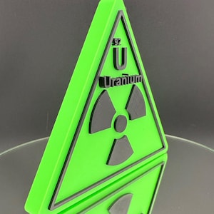 Puede incluir: Un letrero triangular verde neón con un borde negro y un símbolo de radiación. El letrero presenta el símbolo del elemento "U" para Uranio, con el número atómico 92. La palabra "Uranio" está impresa debajo.