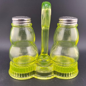 May include: A vintage, light green glass salt and pepper shaker set. The set includes two double-bulb shaped shakers with silver lids, connected by a handle. The base is also light green and has a ribbed design.