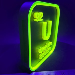 Może przedstawiać: Neonowo-zielony, drukowany w 3D znak z symbolem pierwiastka Uranu, 'U', liczbą atomową '92' i innymi danymi naukowymi. Znak ma czarne tło i jasną zieloną obwódkę, z podświetlonym tekstem i obwódką.