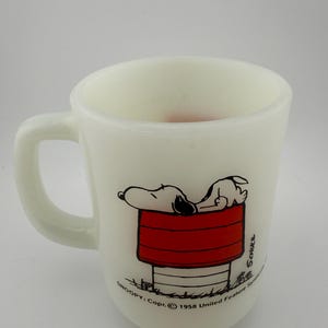 May include: A white milk glass mug featuring a graphic of Snoopy sleeping on top of a red doghouse. The text reads Schulz and SNOOPY: Copr. 1958 United Feature Syndicate, Inc. The mug has a curved handle and a smooth, opaque finish throughout.
