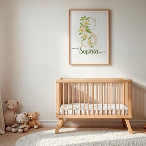 Puede incluir: Una habitación infantil con un estampado enmarcado que muestra el nombre "Sophia" debajo de una letra "S" floral. La habitación cuenta con una cuna de madera, osos de peluche y una alfombra redonda. La combinación de colores es neutra con tonos de madera clara.