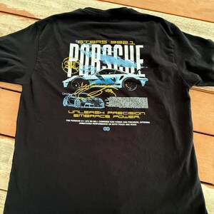 May include: A black t-shirt featuring a graphic on the back with the text PORSCHE, GT3RS 992.1, UNLEASH PRECISION EMBRACE POWER, and a detailed description of the Porsche 911 GT3 RS 992.1, accompanied by illustrations of the sports car.