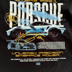 May include: A black t-shirt featuring a graphic of a light blue Porsche 911 GT3 RS 992.1. The design includes the text PORSCHE, GT3 RS 992.1, UNLEASH PRECISION EMBRACE POWER, and a detailed paragraph about the vehicle's performance specifications.