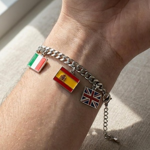 Puede incluir: Pulsera de cadena plateada con colgantes de las banderas italiana, española y británica. Los colgantes cuadrados están rellenos de esmalte. La pulsera se lleva en la muñeca.
