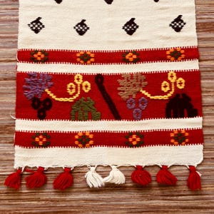Puede incluir: Un tapiz textil tejido rectangular con fondo blanco hueso y motivos de bellotas en marrón oscuro. Incluye bandas horizontales rojas con patrones florales naranjas y verdes, una sección inferior con diseños coloridos de uvas y hojas, y borlas rojas y blancas.