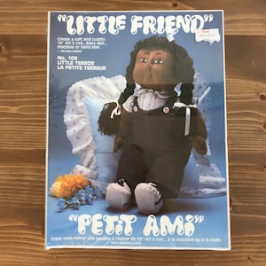 May include: A sewing pattern for a 19-inch (47.5 cm) doll. The doll is black with brown hair and is wearing overalls. The pattern is titled "Little Friend" and "Petit Ami".