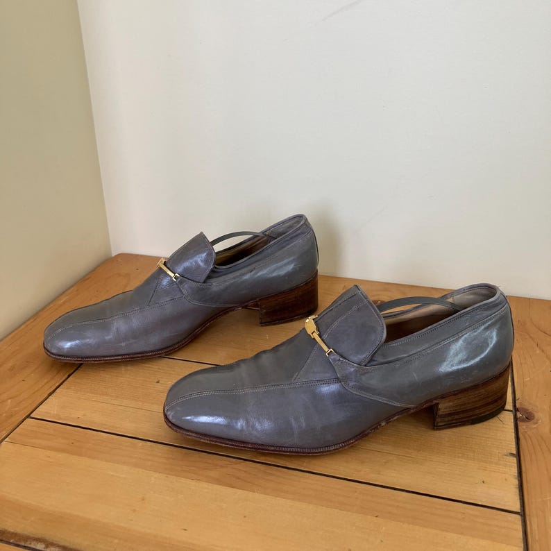 May include: A pair of gray leather loafers with gold buckles. The loafers have a low heel and a pointed toe.