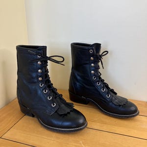 May include: A pair of black leather lace-up boots featuring a rounded toe, a low heel, and a decorative fringe panel covering the base of the laces. The boots have silver-toned metal eyelets and black laces tied at the top of the ankle shaft.