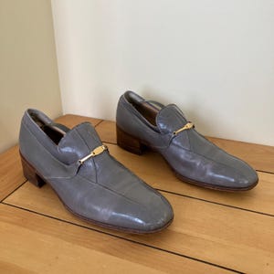 May include: A pair of gray leather slip-on shoes with gold buckles. The shoes have a low heel and a pointed toe.