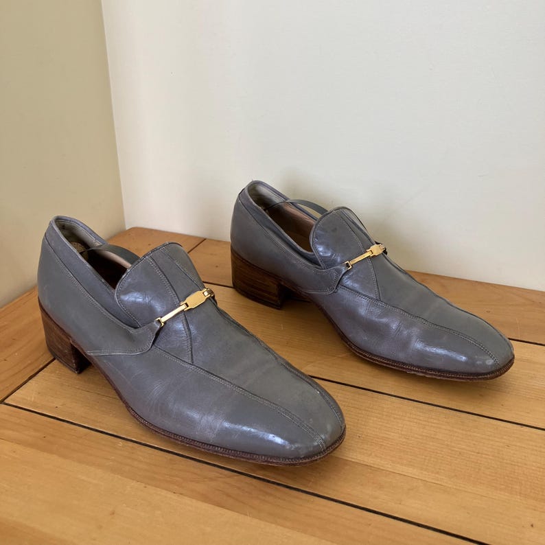 May include: A pair of gray leather slip-on shoes with gold buckles. The shoes have a low heel and a pointed toe.