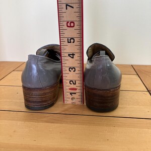 May include: A pair of gray leather loafers with brown heels. The shoes are measured against a ruler, showing they are about 6 inches tall.