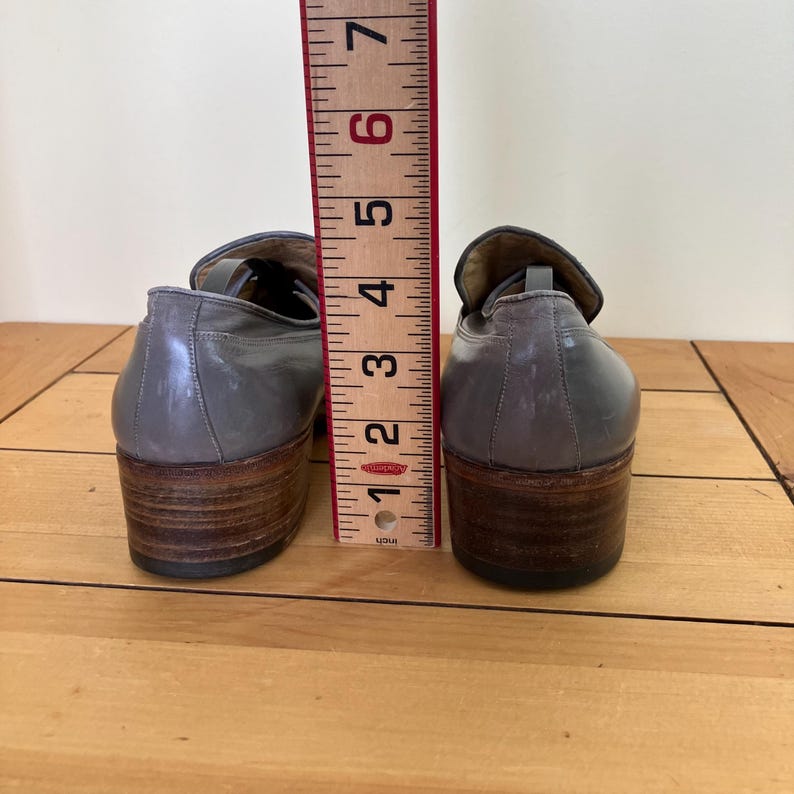 May include: A pair of gray leather loafers with brown heels. The shoes are measured against a ruler, showing they are about 6 inches tall.