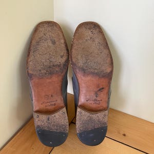May include: Close-up of the soles of a pair of black leather shoes. The shoes are handmade in Italy and have a brown leather sole with the text "Handmade in Italy" printed on them.