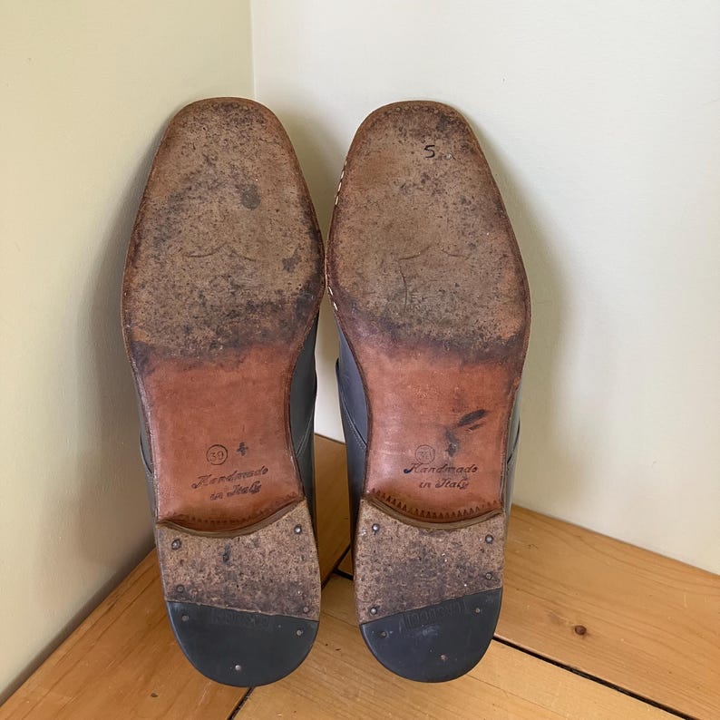 May include: Close-up of the soles of a pair of black leather shoes. The shoes are handmade in Italy and have a brown leather sole with the text "Handmade in Italy" printed on them.