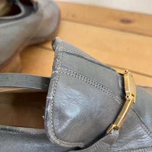 May include: A pair of gray leather loafers with a gold buckle. The loafers have a pointed toe and a low heel.