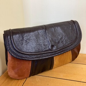 May include: A vintage clutch bag with a flap closure. The bag features a patchwork design with panels of brown, tan, and black leather. The flap is a darker shade of brown with a textured finish. The bag is resting on a wooden surface.