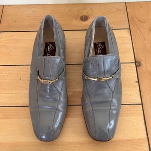 May include: A pair of gray leather slip-on shoes with gold buckles. The shoes have a pointed toe and a low heel. The brand name "Julius" is visible on the inside of the shoes.
