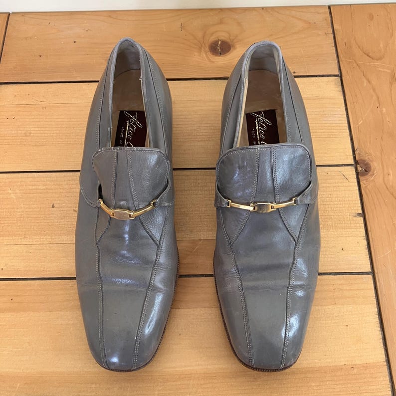 May include: A pair of gray leather slip-on shoes with gold buckles. The shoes have a pointed toe and a low heel. The brand name "Julius" is visible on the inside of the shoes.