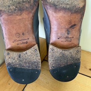 May include: Close-up of the soles of two black leather shoes. The soles are brown leather with the text "Handmade in Italy" and the size "39" and "41" printed on them. The shoes have black rubber soles.