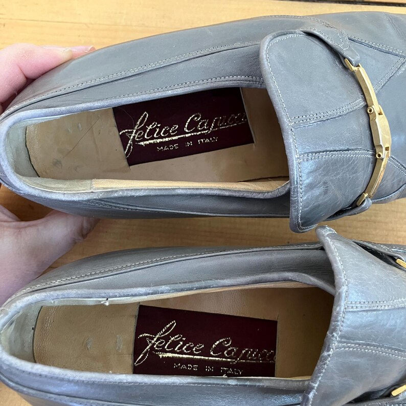 May include: A pair of gray leather loafers with a gold buckle. The loafers have a leather lining and a label that reads "Felice Capucci Made in Italy".