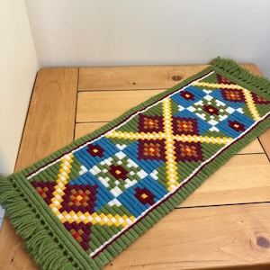 May include: A rectangular, hand-stitched table runner with a geometric pattern. The runner features a green background with a central design in blue, white, yellow, and burgundy. The edges are finished with a green fringe.