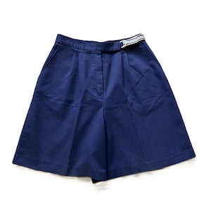 May include: Navy blue shorts with a matching belt. The shorts have a high waist and pleats in the front. The belt is striped with red, white, and blue. The shorts are made of a smooth fabric.